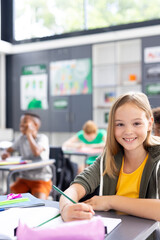 Wall Mural - Vertical portrait of happy caucasian schoolgirl writing at desk in diverse class, copy space