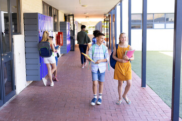 Wall Mural - Smiling caucasian of boy and girl holding books, walking in school corridor talking with copy space