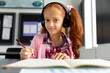 Portrait of happy biracial schoolgirl sitting at desk wearing headphones in class with copy space