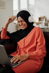 Poster - Young laughing woman in hijab and headphones watching situation comedy on laptop screen while sitting on sofa in front of camera