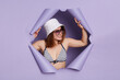 © sementsova321 - Friendly attractive woman wearing swimwear panama and sunglasses breaking through purple paper background looking away with happy face expressing positive emotions.