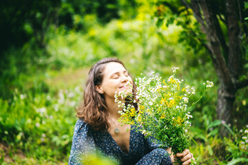 Young cheerful smiling woman in a summer dress picking a bouquet of wild flowers in a summer garden