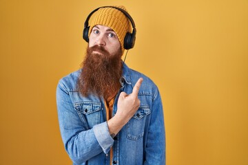 Canvas Print - Caucasian man with long beard listening to music using headphones pointing with hand finger to the side showing advertisement, serious and calm face