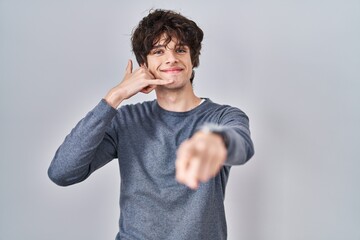 Sticker - Young man standing over isolated background smiling doing talking on the telephone gesture and pointing to you. call me.