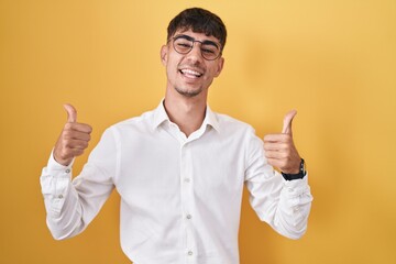 Young hispanic man standing over yellow background success sign doing positive gesture with hand, thumbs up smiling and happy. cheerful expression and winner gesture.