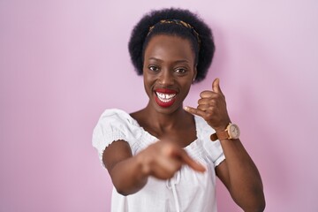 Wall Mural - African woman with curly hair standing over pink background smiling doing talking on the telephone gesture and pointing to you. call me.