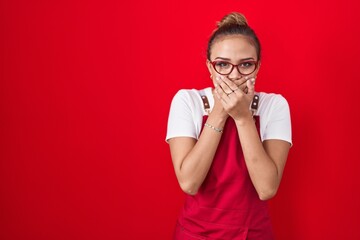 Young hispanic woman wearing waitress apron over red background shocked covering mouth with hands for mistake. secret concept.