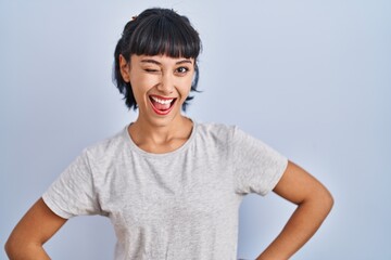 Canvas Print - Young hispanic woman wearing casual t shirt over blue background winking looking at the camera with sexy expression, cheerful and happy face.