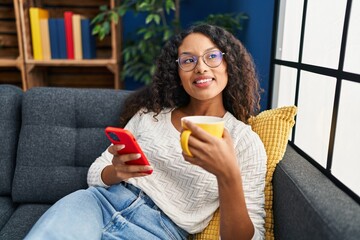 Sticker - Young latin woman drinking coffee and using smartphone at home