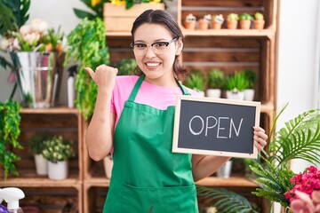 Wall Mural - Hispanic young woman working at florist holding open sign pointing thumb up to the side smiling happy with open mouth