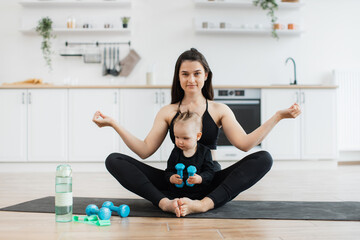 Wall Mural - Full length view of relaxed female sitting in yoga pose with little child on rubber mat in modern apartment. Fit mother exercising with fingers in gyan mudra while cute daughter testing dumbbells.