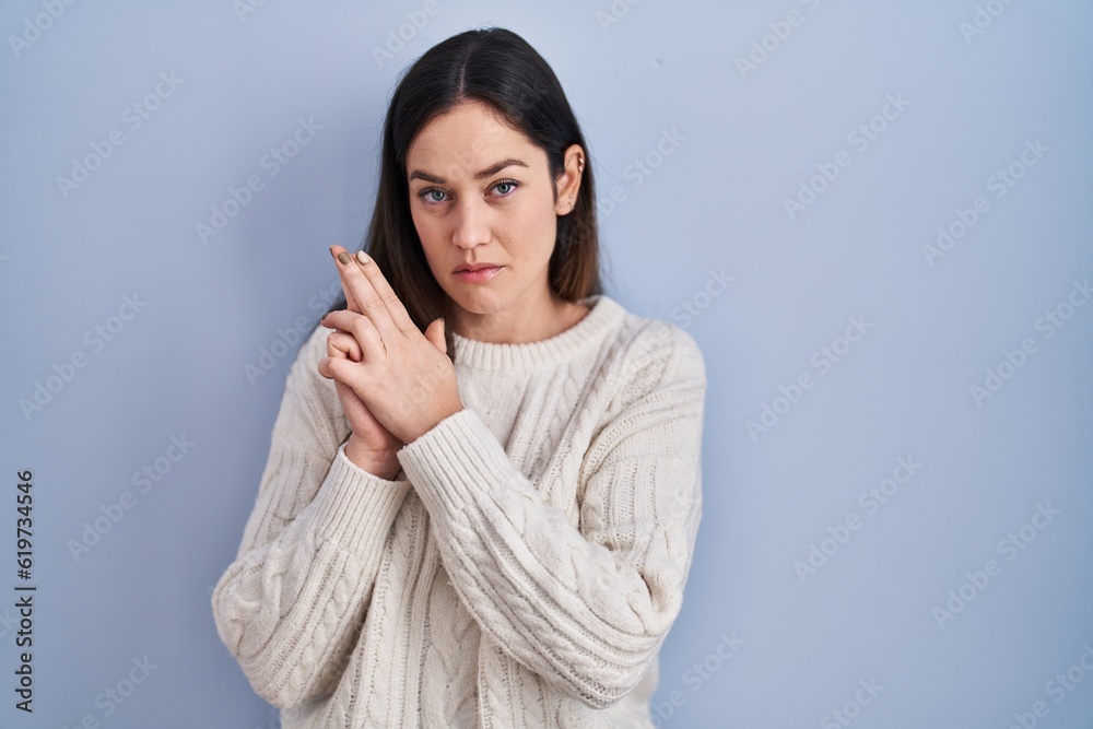 Young brunette woman standing over blue background holding symbolic gun with hand gesture, playing killing shooting weapons, angry face