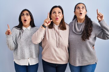 Canvas Print - Mother and two daughters standing over blue background amazed and surprised looking up and pointing with fingers and raised arms.