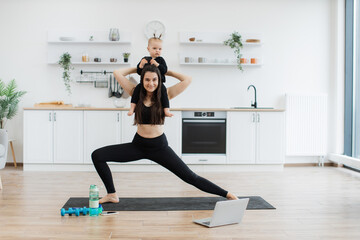 Wall Mural - Sporty female keeping little girl on shoulders while stretching knees in standing position on mat at home. Active mommy in yoga outfit improving fitness while entertaining daughter at weekend.