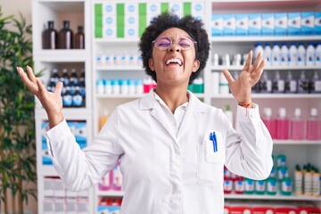 Wall Mural - Beautiful african woman with curly hair working at pharmacy drugstore celebrating mad and crazy for success with arms raised and closed eyes screaming excited. winner concept