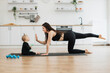 Side view of beautiful woman giving high five to cute baby girl while achieving yoga position indoors. Athletic mother in sports clothes turning every day workout into play on kitchen background.