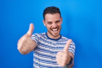 Canvas Print - Young hispanic man standing over blue background approving doing positive gesture with hand, thumbs up smiling and happy for success. winner gesture.