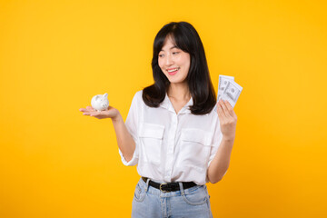 Poster - Portrait beautiful young business asian woman wearing white shirt and denim jean with a lot of cash money and piggy bank isolated on yellow background. Wealth money saving, finance investment concept.
