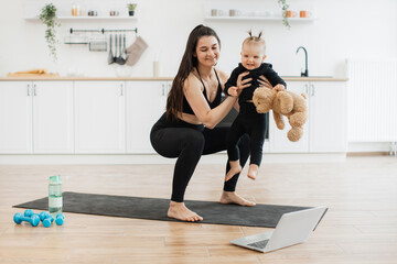 Wall Mural - Smiling female holding cute girl with soft toy while working out squats to online video on kitchen background. Athletic mommy and child in yoga outfits enjoying benefits of quality time together.