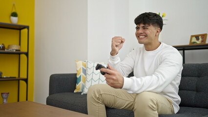 Poster - Young hispanic man playing video game sitting on sofa with winner expression at home