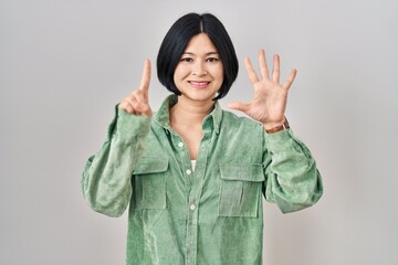 Sticker - Young asian woman standing over white background showing and pointing up with fingers number six while smiling confident and happy.