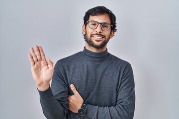 Wall Mural - Handsome latin man standing over isolated background waiving saying hello happy and smiling, friendly welcome gesture