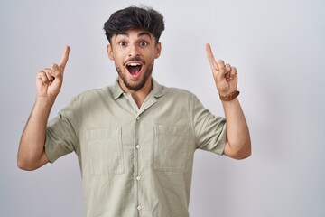 Poster - Arab man with beard standing over white background smiling amazed and surprised and pointing up with fingers and raised arms.