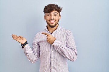 Sticker - Arab man with beard standing over blue background amazed and smiling to the camera while presenting with hand and pointing with finger.