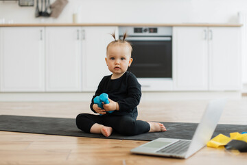 Wall Mural - Full length portrait of sweet baby girl in black outfit sitting on yoga mat with blue resistance band in hand indoors. Adorable little infant being introduced to physical activities in home interior.