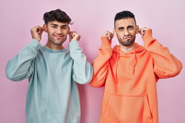 Wall Mural - Young hispanic gay couple standing over pink background smiling pulling ears with fingers, funny gesture. audition problem