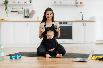 Wall Mural - Portrait of smiling family woman helping sweet daughter with lifting dumbbells on black mat in kitchen. Cheerful female parent encouraging healthy habits while enhancing kid's strength at home.