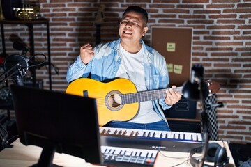 Wall Mural - Hispanic young man playing classic guitar at music studio very happy and excited doing winner gesture with arms raised, smiling and screaming for success. celebration concept.