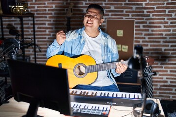 Poster - Hispanic young man playing classic guitar at music studio very happy and excited doing winner gesture with arms raised, smiling and screaming for success. celebration concept.