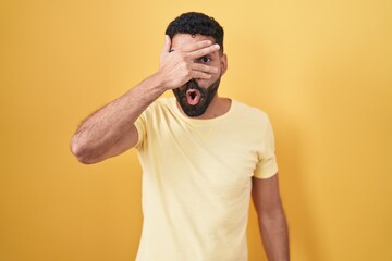 Canvas Print - Hispanic man with beard standing over yellow background peeking in shock covering face and eyes with hand, looking through fingers with embarrassed expression.