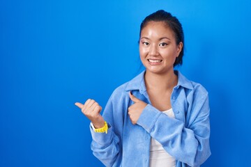 Sticker - Asian young woman standing over blue background pointing to the back behind with hand and thumbs up, smiling confident