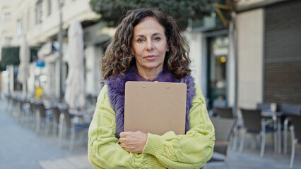 Sticker - Middle age hispanic woman standing with serious expression holding clipboard at coffee shop terrace