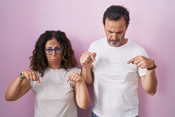 Sticker - Middle age hispanic couple together over pink background pointing down looking sad and upset, indicating direction with fingers, unhappy and depressed.