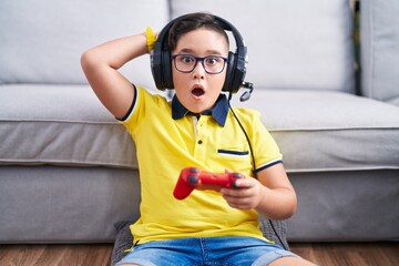 Canvas Print - Young hispanic kid playing video game holding controller wearing headphones crazy and scared with hands on head, afraid and surprised of shock with open mouth