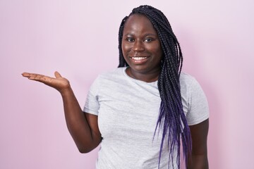 Sticker - Young african woman standing over pink background smiling cheerful presenting and pointing with palm of hand looking at the camera.