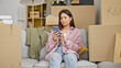 © Krakenimages.com - Young beautiful hispanic woman using smartphone sitting on sofa at new home