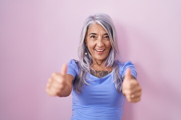 Wall Mural - Middle age woman with tattoos standing over pink background approving doing positive gesture with hand, thumbs up smiling and happy for success. winner gesture.