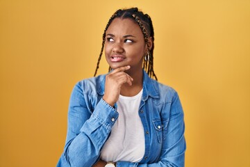 Wall Mural - African american woman with braids standing over yellow background thinking worried about a question, concerned and nervous with hand on chin