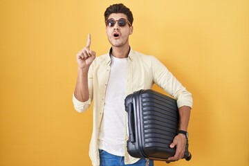 Wall Mural - Young hispanic man holding suitcase going on summer vacation amazed and surprised looking up and pointing with fingers and raised arms.