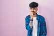 Young hispanic man with beard standing over pink background looking stressed and nervous with hands on mouth biting nails. anxiety problem.