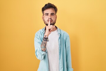 Poster - Young hispanic man with tattoos standing over yellow background asking to be quiet with finger on lips. silence and secret concept.