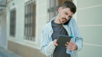 Sticker - Young hispanic man using touchpad talking on smartphone holding bottle of water at street