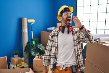 Wall Mural - Young hispanic man with beard working at home renovation shouting and screaming loud to side with hand on mouth. communication concept.