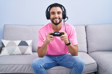 Wall Mural - Hispanic young man playing video game holding controller sitting on the sofa with a happy and cool smile on face. lucky person.