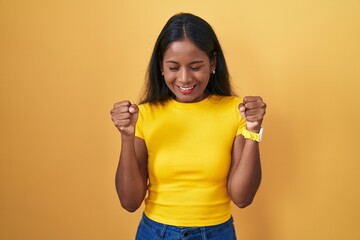 Young indian woman standing over yellow background excited for success with arms raised and eyes closed celebrating victory smiling. winner concept.