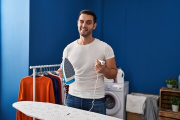 Poster - Young hispanic man with beard ironing clothes at home winking looking at the camera with sexy expression, cheerful and happy face.